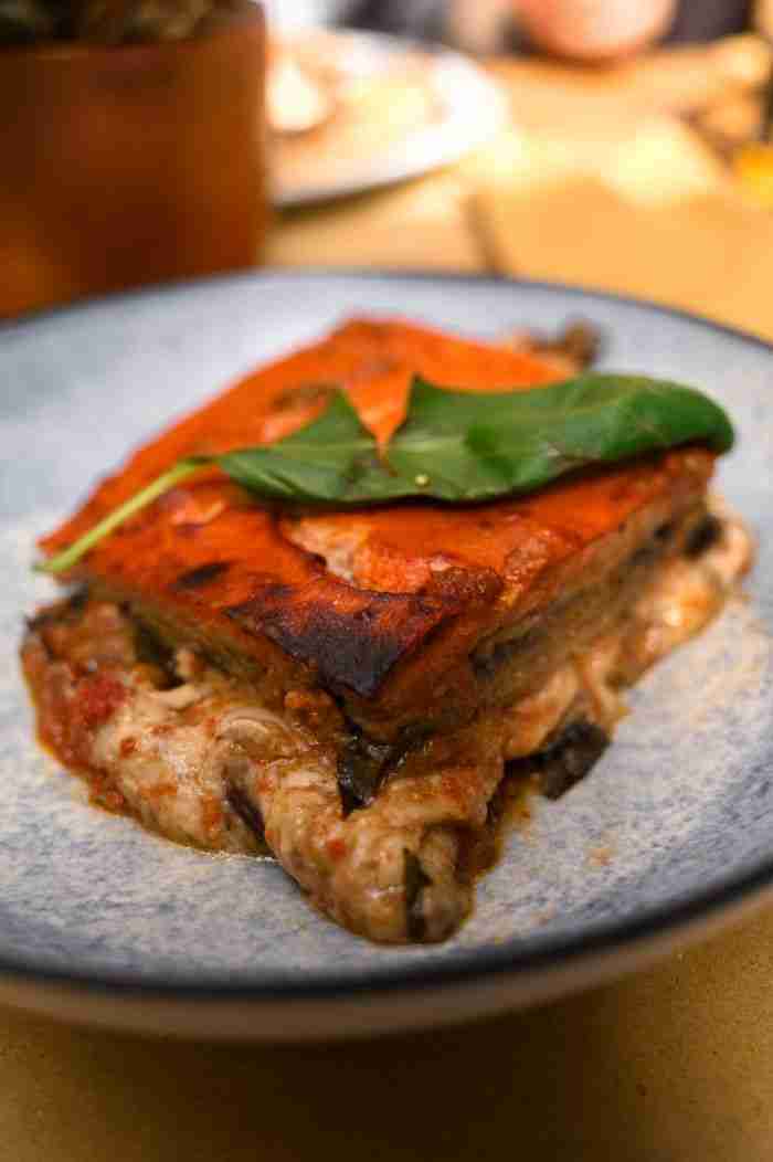 contact-img Close-up of a savory eggplant lasagna slice topped with a basil leaf, served on a plate.