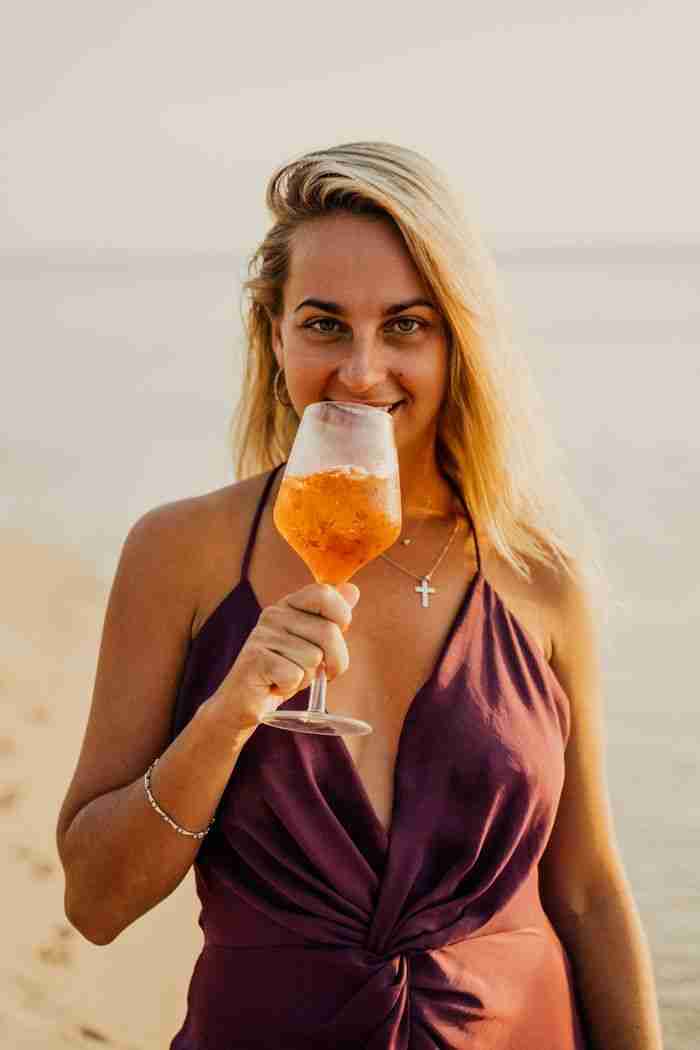 Woman stands on the sandy beach holding a cocktail, smiling warmly at sunset.